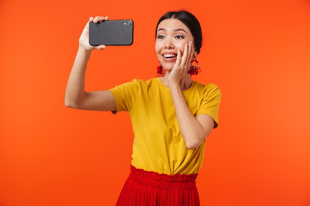 bella giovane donna felice che posa isolata sopra la parete arancione che parla dal telefono cellulare prende un selfie.