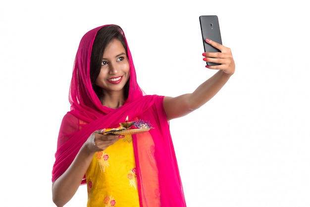 Bella giovane donna felice che prende selfie con il thali di pooja facendo uso di un telefono cellulare o di uno smartphone su bianco