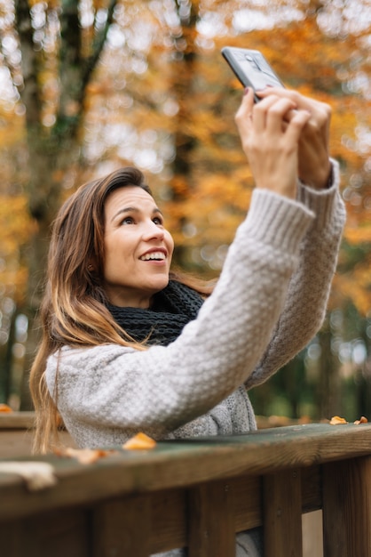 Bella giovane donna felice che prende selfie con lo smartphone nel parco di autunno. Concetto di stagione, tecnologia e persone.