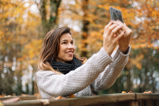 Bella giovane donna felice che prende selfie con lo smartphone nel parco di autunno. Concetto di stagione, tecnologia e persone.