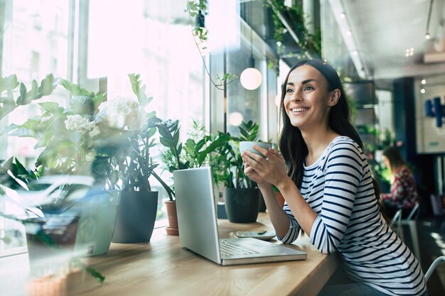 Bella giovane donna felice che usa il computer portatile e beve una tazza di caffè nella caffetteria durante l'ora di pranzo