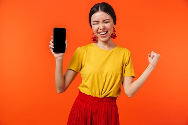 bella giovane donna felice emozionante che posa isolata sopra la parete arancione che mostra il display del telefono cellulare.