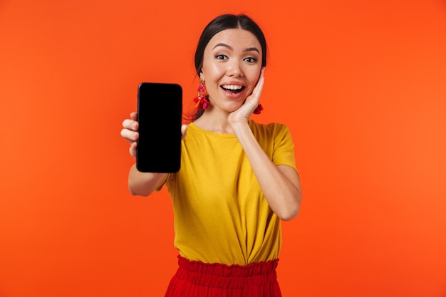 bella giovane donna felice emozionante che posa isolata sopra la parete arancione che mostra il display del telefono cellulare.