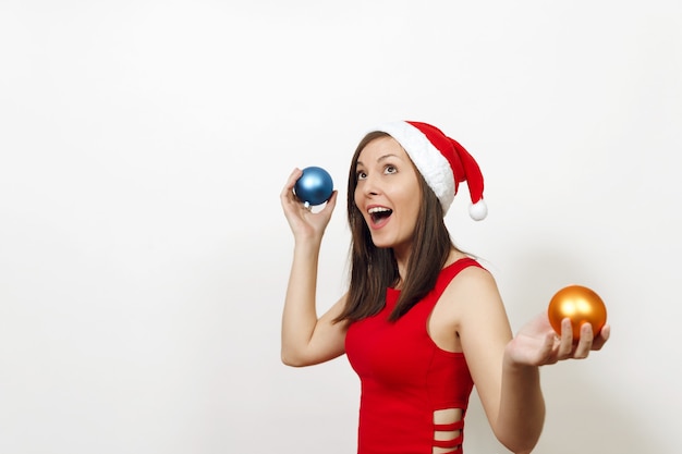 Bella giovane donna felice europea con pelle sana e sorriso affascinante in vestito rosso e cappello di Natale che tiene due giocattoli dell'albero su priorità bassa bianca. Ragazza di Santa isolata. Concetto di vacanza di Capodanno 2018.