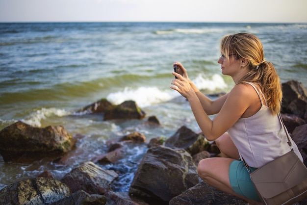 Bella giovane donna felice in abiti leggeri e leggeri con una treccia attenta scatta foto del paesaggio dell'orizzonte marino su un telefono moderno mentre è seduto su pietre bagnate