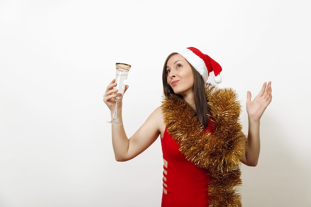 Bella giovane donna felice sorridente caucasica in vestito rosso, cappello di Natale e ghirlanda di orpelli in piedi con un bicchiere di champagne su sfondo bianco. Ragazza di Santa isolata. Concetto di vacanza di Capodanno.