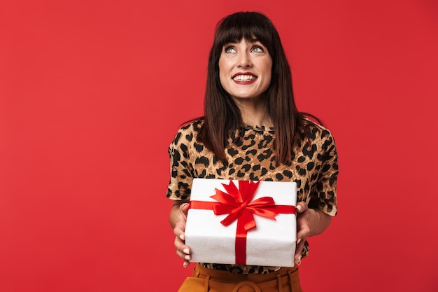 bella giovane donna felice vestita di camicia stampata animale in posa isolata sul muro rosso che tiene la scatola presente.