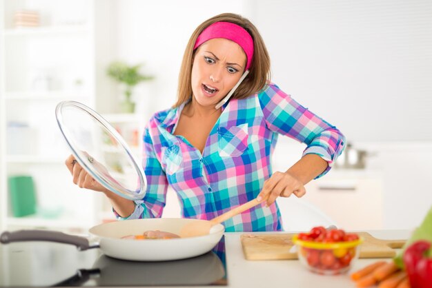 Bella giovane donna furiosa che prepara un pasto sano e usa il telefono nella cucina domestica.