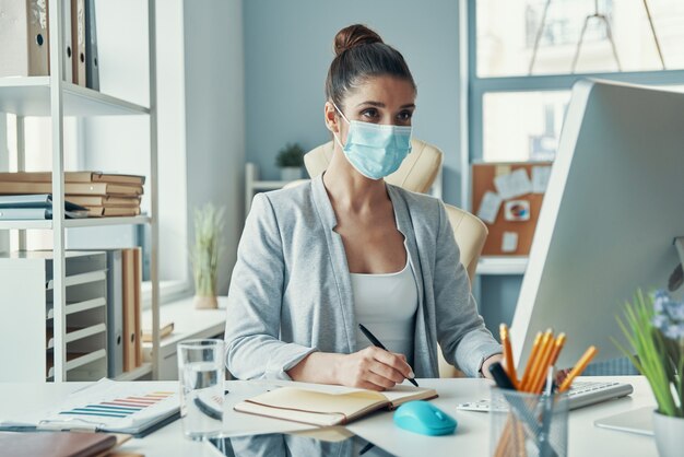 Bella giovane donna in abbigliamento casual intelligente che scrive qualcosa mentre è seduta in ufficio