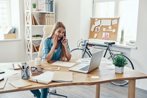 Bella giovane donna in abbigliamento casual intelligente che sorride e parla al telefono mentre è seduta in ufficio