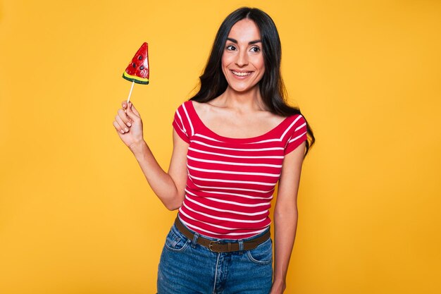 Bella giovane donna in abiti casual con lecca-lecca in mano in posa su sfondo arancione