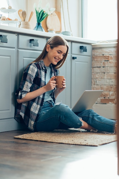 Bella giovane donna in abiti casual sul pavimento della cucina usando un computer portatile e bevendo tè