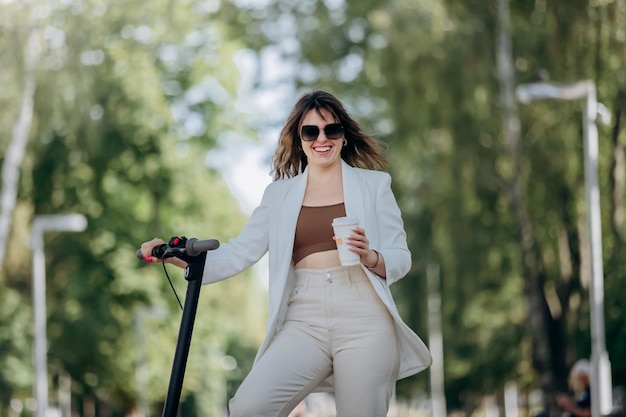 Bella giovane donna in abito bianco e occhiali da sole in piedi con il suo scooter elettrico nel parco cittadino e bere caffè