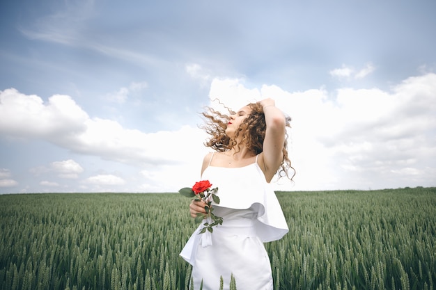 Bella giovane donna in abito bianco in piedi nel campo con rosa in mano