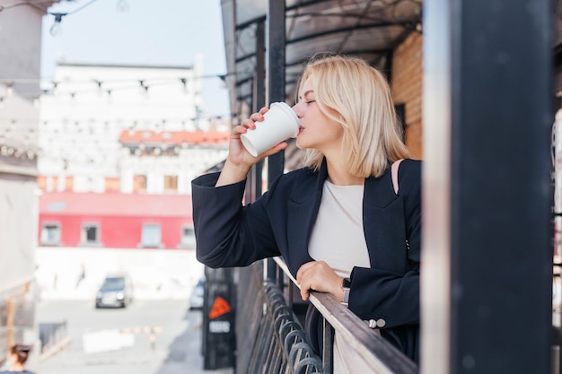 Bella giovane donna in abito e giacca cammina per la città e beve caffè