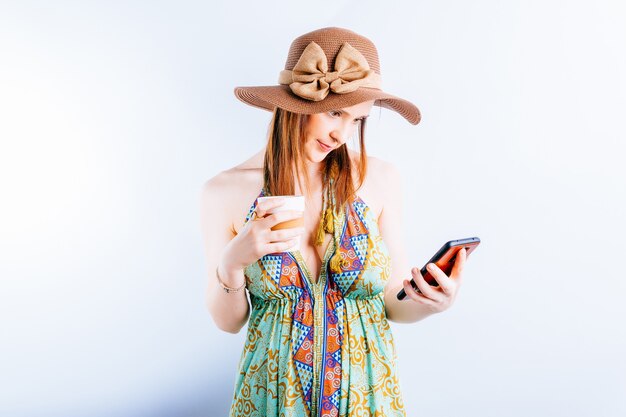 Bella giovane donna in abito estivo, cappello guardando lo smartphone con caffè o tè. concetto di vacanza estiva. connettività