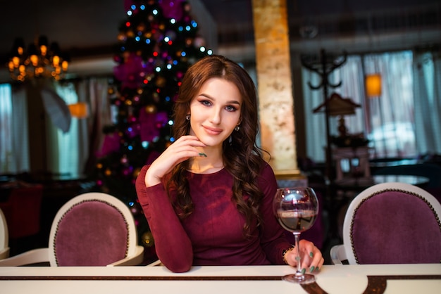 Bella giovane donna in abito grigio sta con un bicchiere di vino in mano su sfondo di albero di Natale, luci e ghirlande.