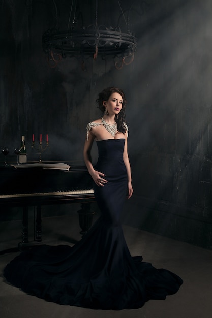Bella giovane donna in abito nero accanto a un pianoforte con candele candelabri e vino, atmosfera drammatica scuro del castello. Boemia.