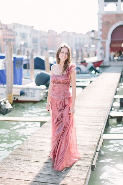 Bella giovane donna in abito rosa in piedi sullo sfondo del canale di Venezia, edifici e gondola. Italia, Europa