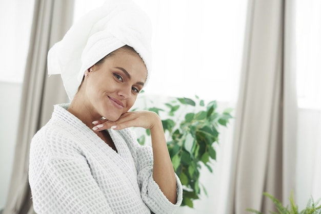 Bella giovane donna in accappatoio e asciugamano sulla testa sorridendo alla telecamera in bagno.