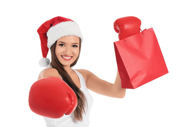 Bella giovane donna in cappello di Babbo Natale con guantoni da boxe e borsa della spesa su sfondo bianco. Concetto di Santo Stefano