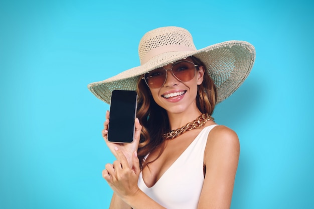 Bella giovane donna in cappello elegante che mostra il suo telefono cellulare mentre sta in piedi su sfondo blu