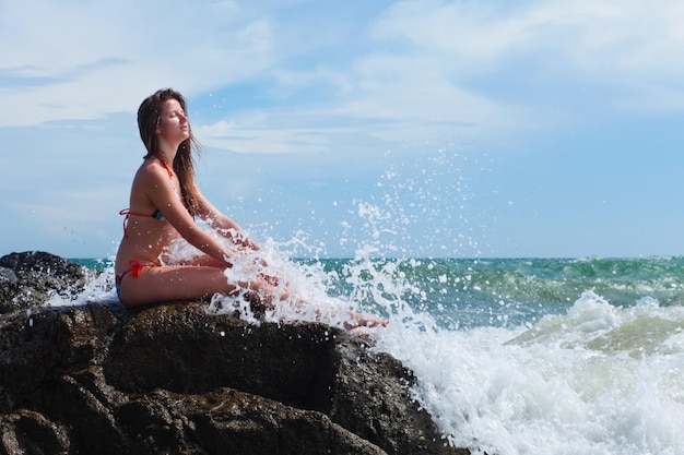 Bella giovane donna in costume da bagno seduta su una pietra contro l'onda e il cielo blu