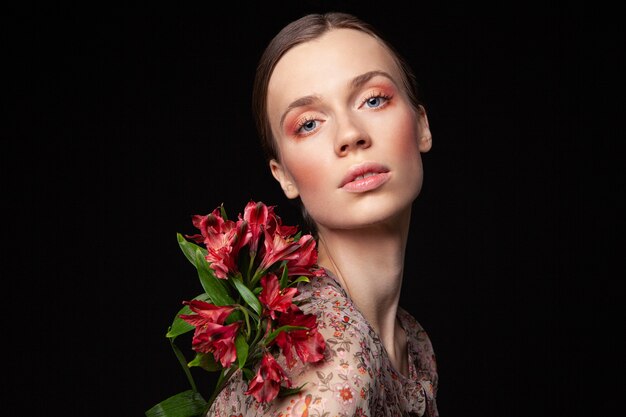 Bella giovane donna in elegante camicetta rossa che tocca il viso con fiori naturali e guarda lontano su sfondo nero