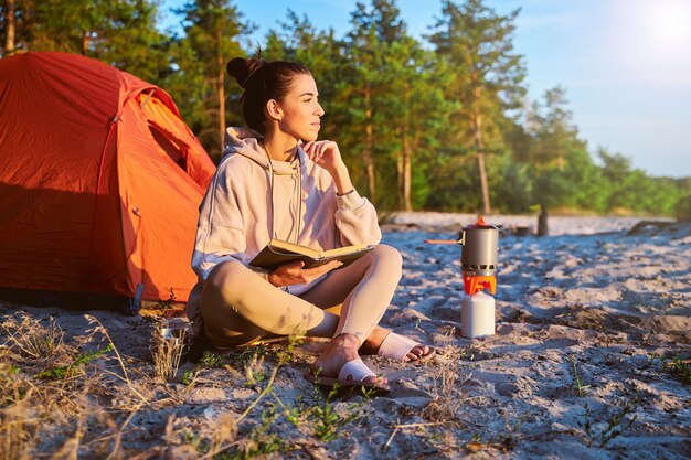 Bella giovane donna in felpa che tiene un libro e sorride mentre è seduta sulla sabbia vicino alla tenda del campo camp