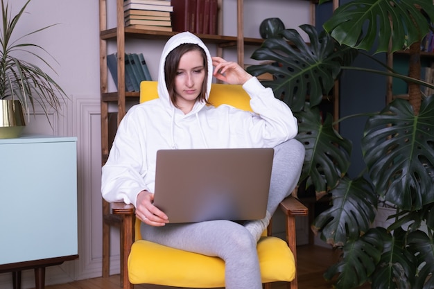 Bella giovane donna in felpa con cappuccio bianca utilizzando laptop seduto sulla poltrona gialla a casa