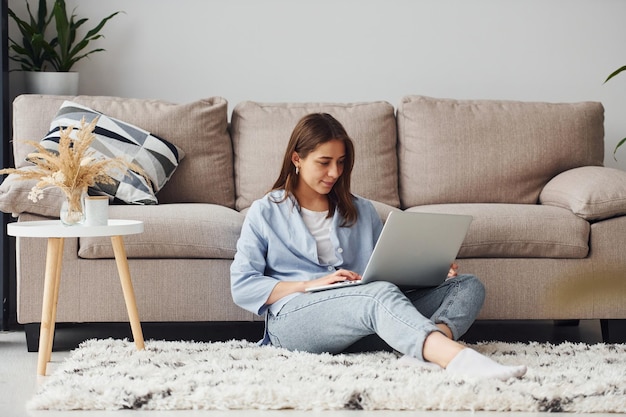 Bella giovane donna in jeans e camicia blu si siede sul pavimento con il computer portatile al chiuso a casa