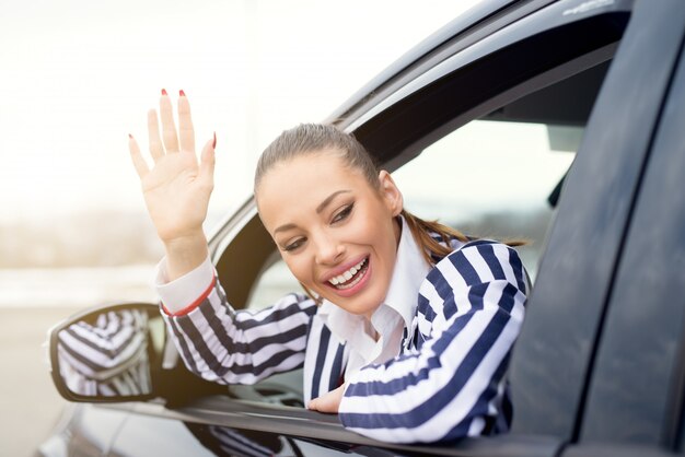 Bella giovane donna in macchina, salutando fuori dalla finestra e sorridente.