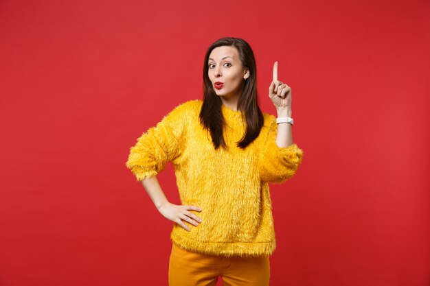 Bella giovane donna in maglione di pelliccia gialla che tiene il dito indice in alto con una grande nuova idea isolata su sfondo rosso brillante della parete in studio. Persone sincere emozioni, concetto di stile di vita. Mock up copia spazio.