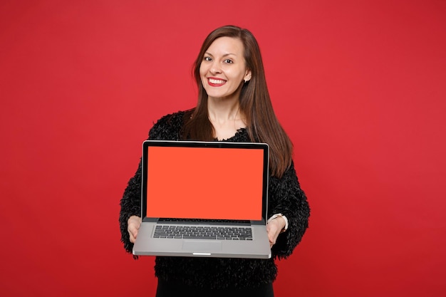 Bella giovane donna in maglione di pelliccia nera che tiene computer pc portatile con schermo vuoto vuoto isolato su sfondo muro rosso in studio. Persone sincere emozioni, concetto di stile di vita. Mock up copia spazio.