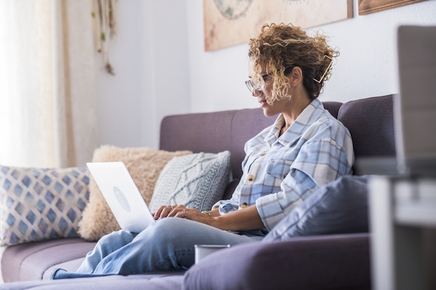 Bella giovane donna in occhiali che lavora al computer portatile che si siede sul sofà. Imprenditrice che lavora da casa. Giovane donna casual seduta sul divano. Donna rilassata che usa il portatile sul divano di casa