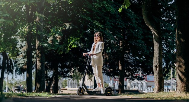 Bella giovane donna in occhiali da sole e abito bianco è in piedi con il suo scooter elettrico nel parco cittadino