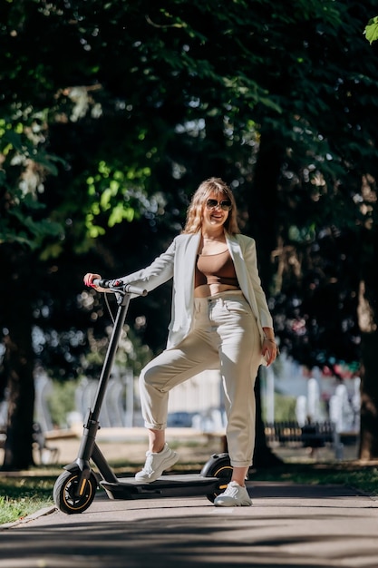 Bella giovane donna in occhiali da sole e abito bianco è in piedi con il suo scooter elettrico nel parco cittadino