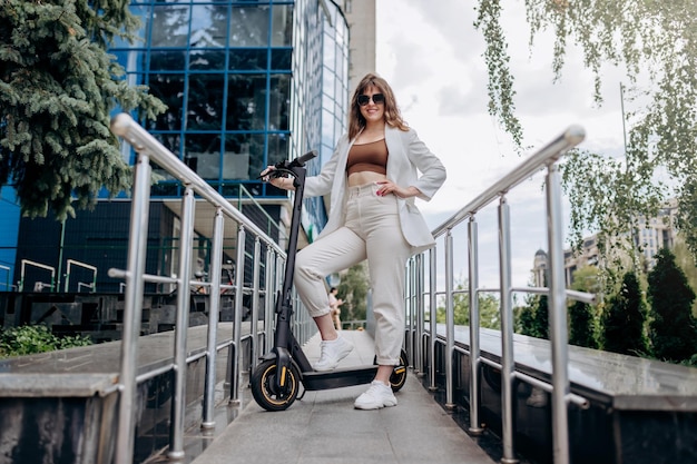 Bella giovane donna in occhiali da sole e abito bianco in piedi con il suo scooter elettrico vicino a un edificio moderno e guardando la fotocamera