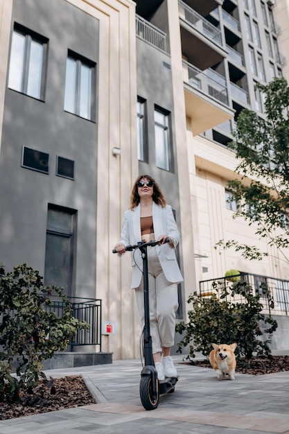 Bella giovane donna in occhiali da sole e abito bianco sta cavalcando sul suo scooter elettrico con il cane Corgi vicino a un edificio moderno in città e distoglie lo sguardo