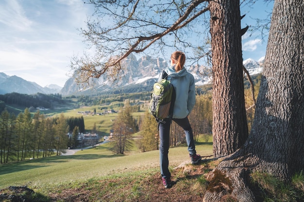 Bella giovane donna in occhiali da sole e giacca blu è in piedi sulla collina sotto l'albero al tramonto Colorato paesaggio primaverile con ragazza sportiva prati alberi verdi e montagne Viaggio in Italia