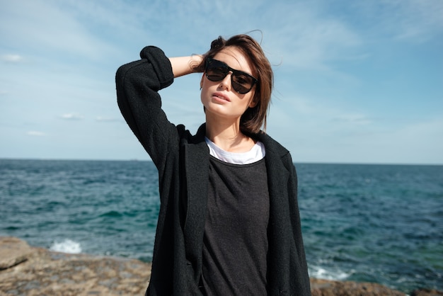 Bella giovane donna in occhiali da sole in piedi in riva al mare