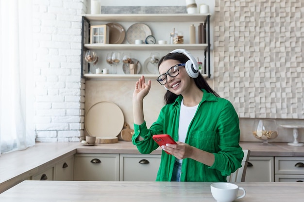 Bella giovane donna in occhiali e camicia verde che si rilassa a casa in cucina ascoltando musica dentro