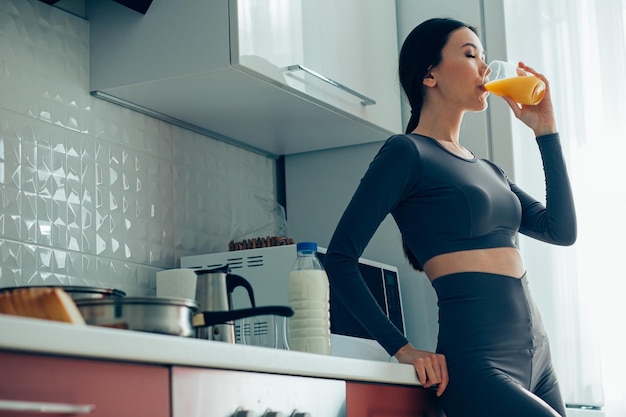 Bella giovane donna in piedi in cucina con un bicchiere di succo d'arancia e bevendolo con gli occhi