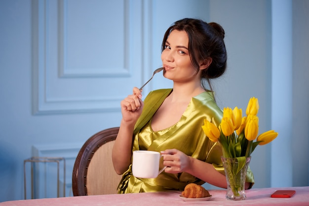 Bella giovane donna in pigiama bere caffè al mattino con croissant.