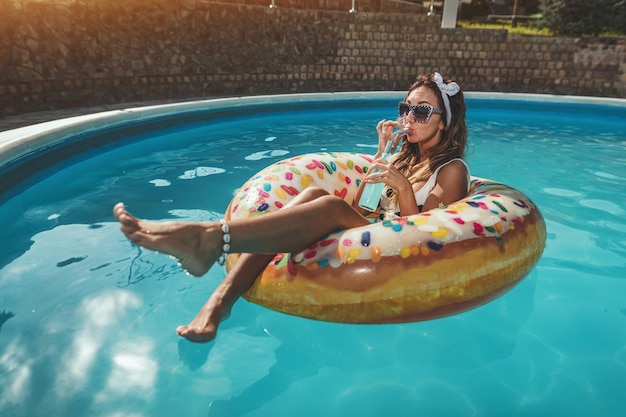 Bella giovane donna in piscina nuota sulla ciambella gonfiabile e si diverte con un bicchiere di cocktail in vacanza.