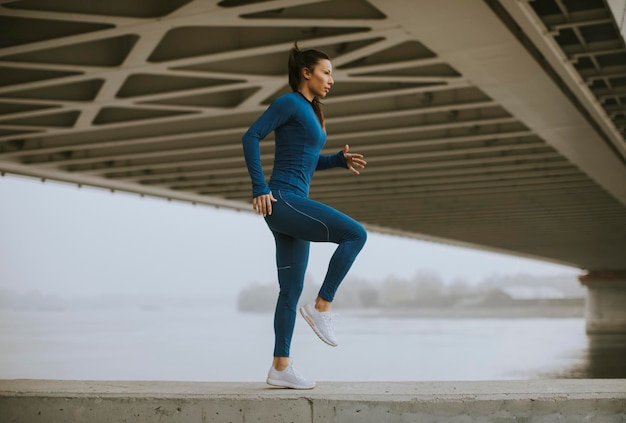 Bella giovane donna in tuta blu che si allunga prima dell'allenamento in riva al fiume al mattino d'autunno