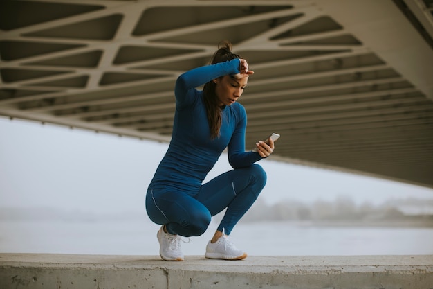 Bella giovane donna in tuta da ginnastica blu che monitora i suoi progressi di allenamento sull'app di fitness vicino al fiume al mattino d'autunno