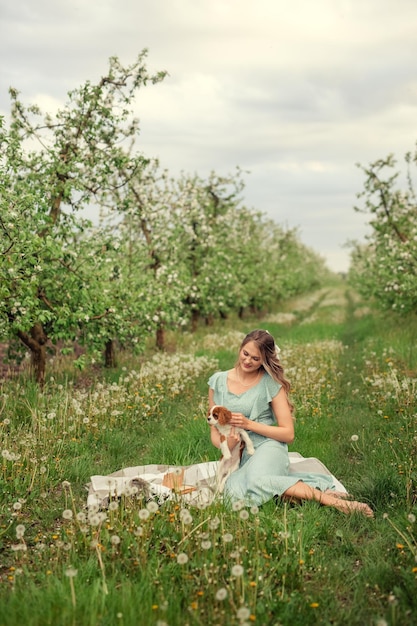 Bella giovane donna in un abito romantico su un picnic in primavera in un giardino fiorito con un cucciolo