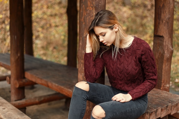 Bella giovane donna in un maglione lavorato a maglia alla moda si siede su una panca di legno in natura