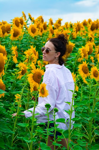 Bella giovane donna in una camicia bianca e occhiali da sole in posa tra girasoli gialli in una giornata estiva.
